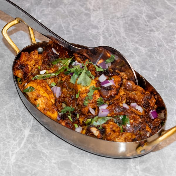 Indian chicken dish in a brass-handled serving bowl, garnished with red onions and cilantro.