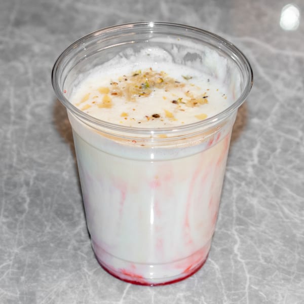 A clear plastic cup of Indian lassi, white with red swirls, topped with nuts and spices.