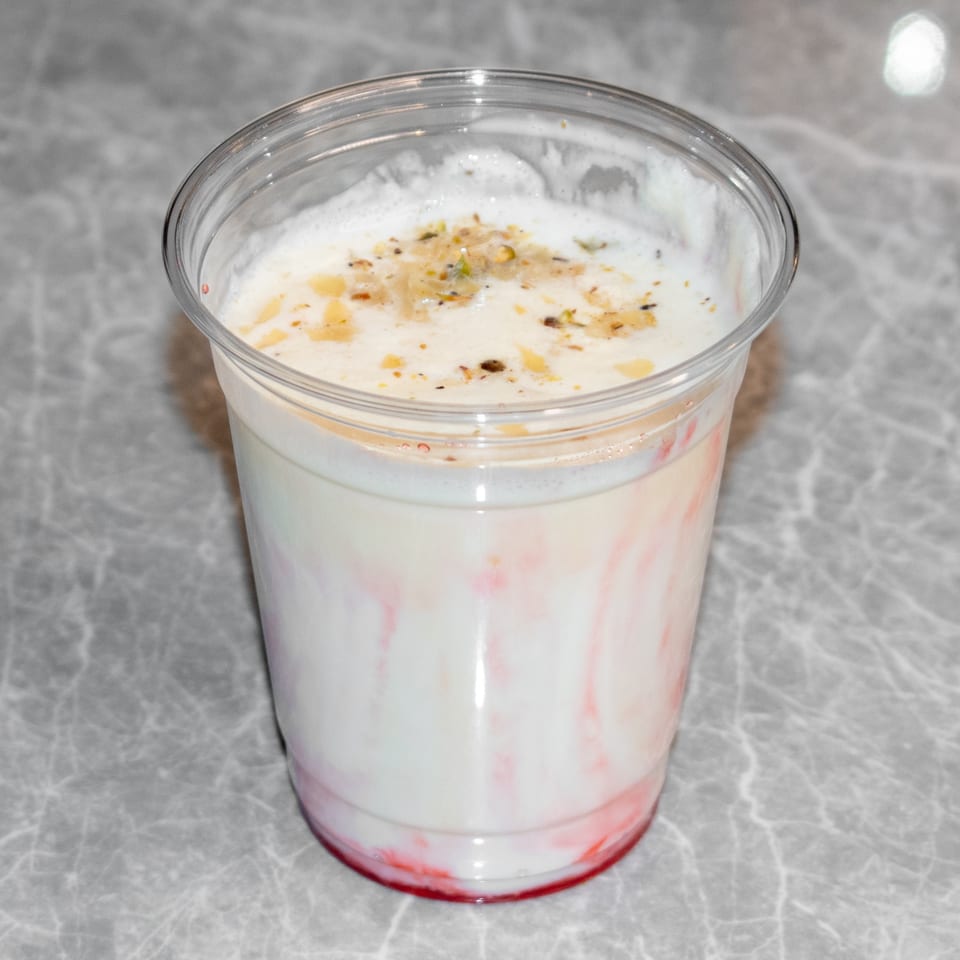 A clear plastic cup of Indian lassi, white with red swirls, topped with nuts and spices.