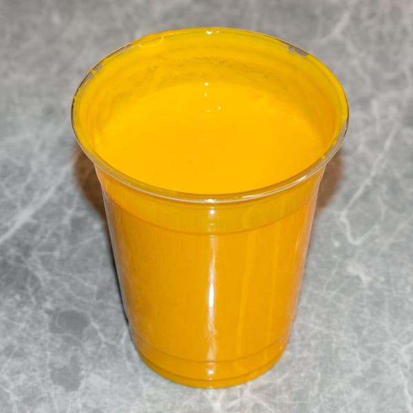 A vibrant yellow mango lassi in a clear plastic cup, sitting on a gray marble surface.