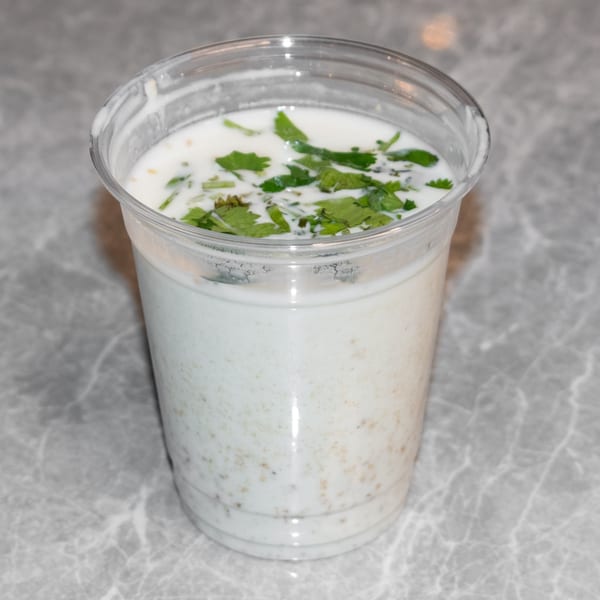 A clear plastic cup of white Indian lassi, topped with fresh green cilantro, on a gray marble surface.